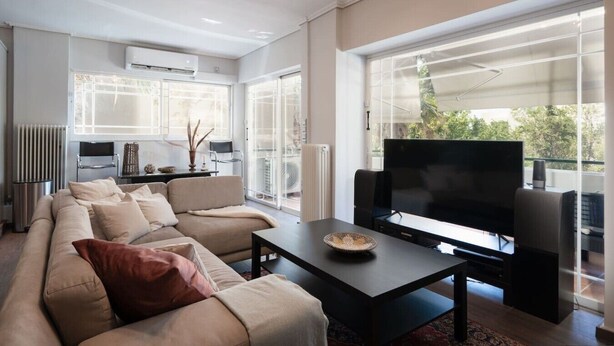 A modern living room with a sofa, a coffee table, and a flat-screen TV. There is a large window providing a view of the outdoors. - Luxe Bliss – 2BR, Glyfada (Glifada)