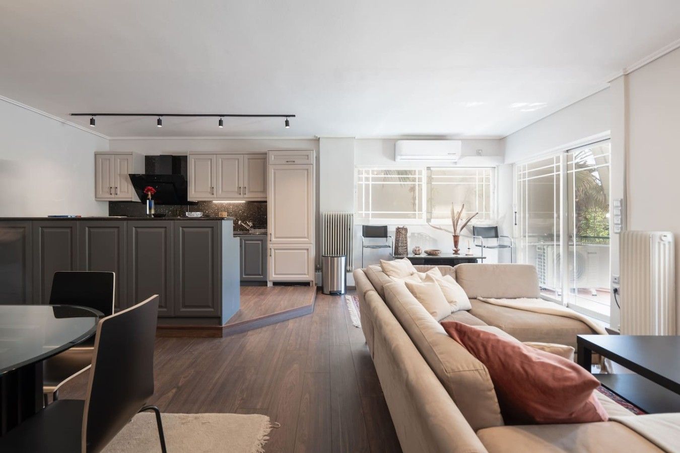 A modern kitchen with grey cabinets, a dining area with a round table and chairs, and a living room with a beige sofa and large windows.
