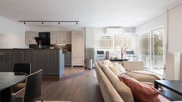 A modern kitchen with grey cabinets, a dining area with a round table and chairs, and a living room with a beige sofa and large windows.