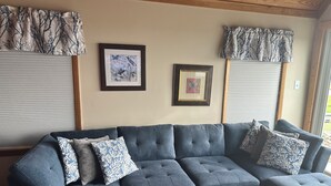 A sectional sofa with multiple cushions, a framed butterfly artwork, and a window with a patterned valance. - Tranquil 1-bedroom house with AC on charming Smith Island (Smith Island)