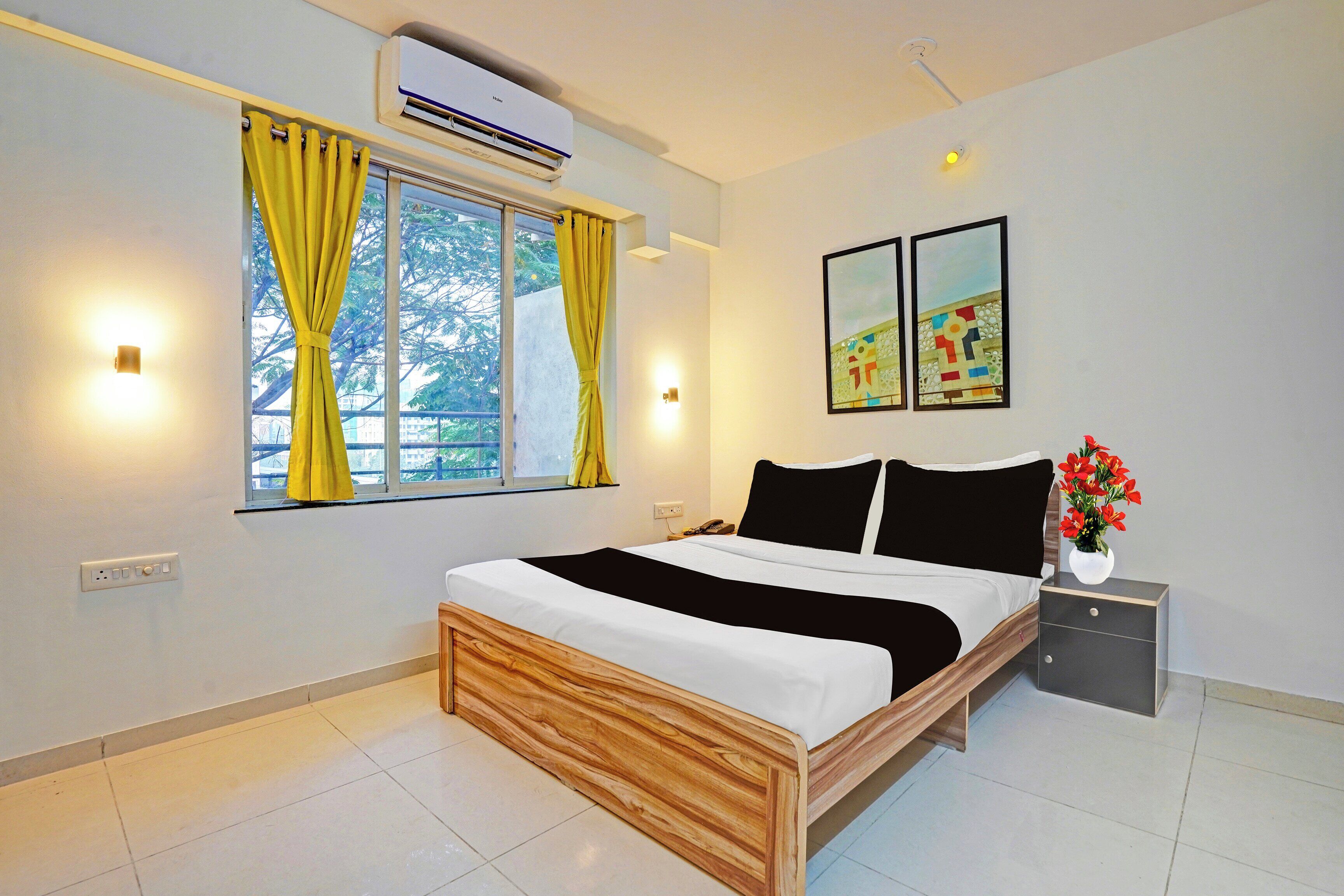A hotel room with a bed, a bedside table, a nightstand, a wall-mounted air conditioner, a window with curtains, and two framed pictures on the wall.