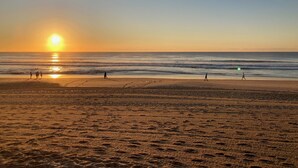 Una playa con huellas que conducen al agua, cielo despejado y el sol poniéndose en el horizonte.