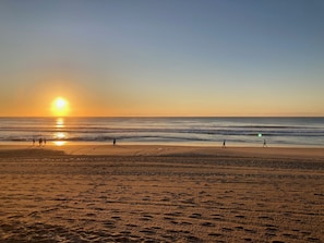 A beach with footprints leading to the water, a clear sky, and the sun setting over the horizon.