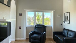 Una sala de estar con un sillón y un sofá de cuero negro, un ventilador de techo y un ventanal con vistas a la vegetación.