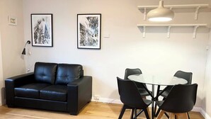 Una sala moderna con un sofá de cuero negro, una mesa de comedor con tapa de vidrio y cuatro sillas, y dos cuadros abstractos en la pared.