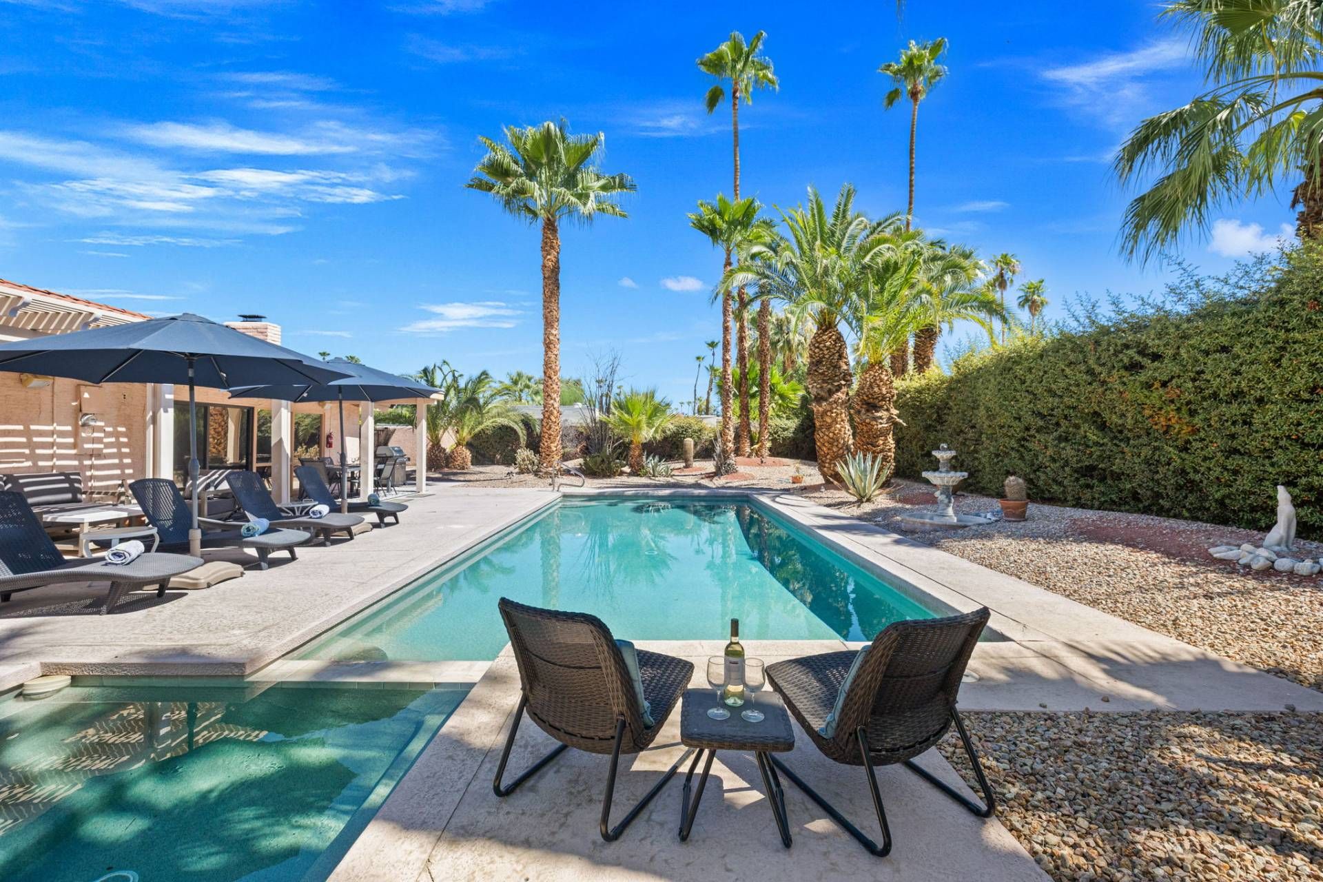A swimming pool with lounge chairs and umbrellas, surrounded by palm trees and a hedge.