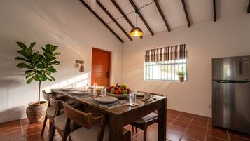 A dining area with a wooden table set for four, a potted plant, a fruit bowl, and a refrigerator.