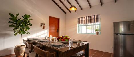 Un comedor con una mesa de madera dispuesta para cuatro, una planta en maceta, un frutero y un refrigerador.