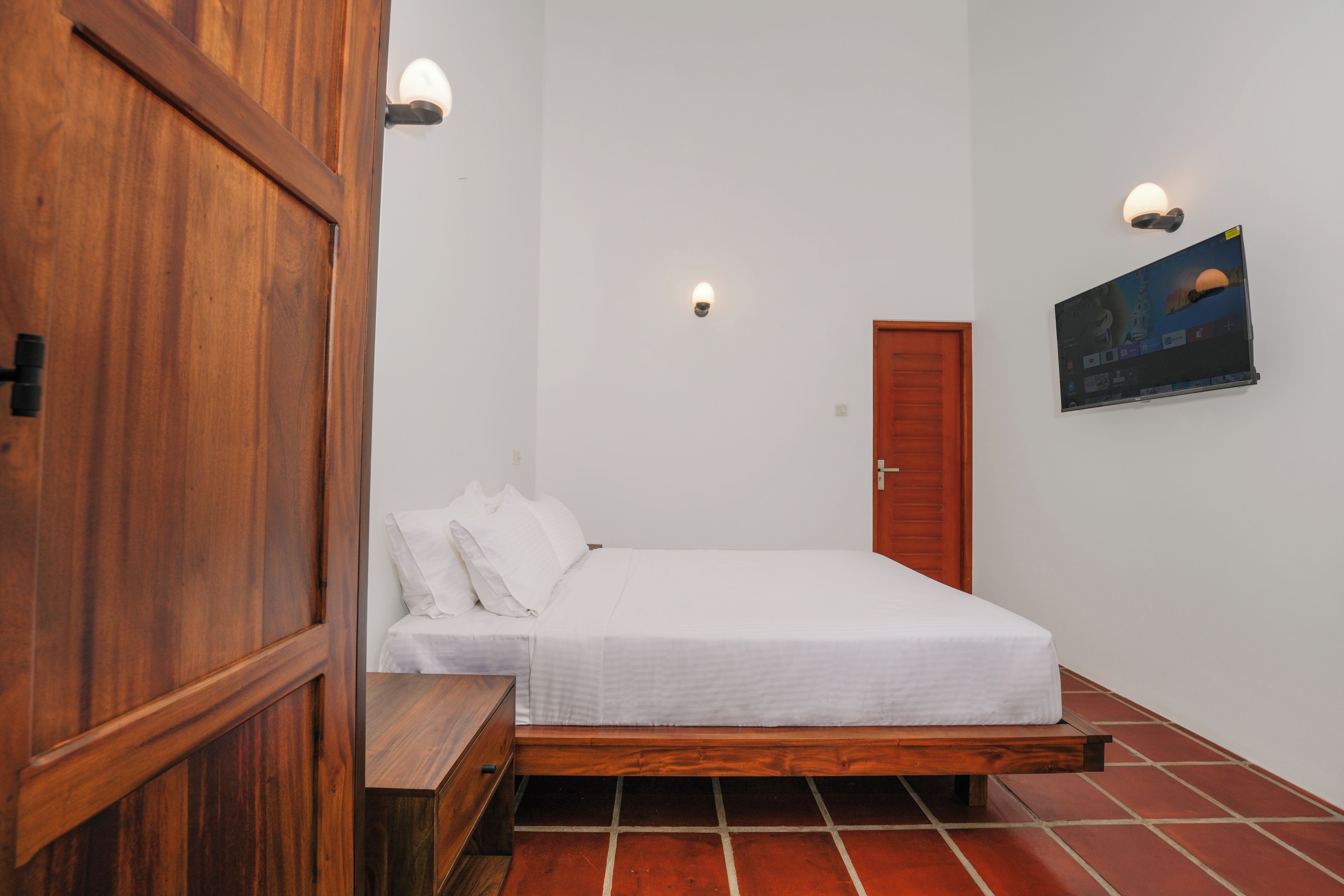 Ein Schlafzimmer mit hölzernem Bettgestell, weißer Bettwäsche, einer Holzbank, einem an der Wand montierten Fernseher und einer Tür.