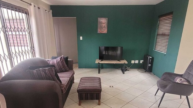 Un soggiorno con divano, poggiapiedi e poltrona, una televisione su un mobile basso e un altoparlante.