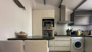 Een compacte keuken met een magnetron, oven, broodrooster en wasmachine. Er is een eettafel met stoelen en een mand met brood op het aanrecht.