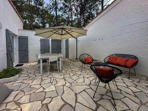Eine Terrasse mit Steinboden, einem weißen Tisch mit Stühlen, einem beigen Sonnenschirm und zwei schwarzen Stühlen mit roten Kissen.