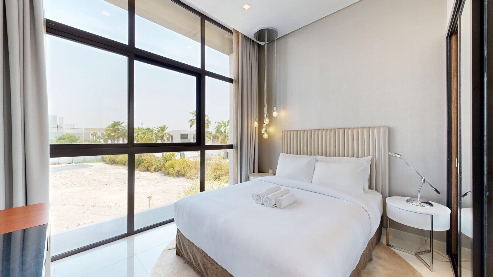 Une chambre d’hôtel moderne dotée d’un grand lit, d’une table de chevet avec une lampe et offrant une vue sur l’extérieur grâce à de grandes fenêtres.