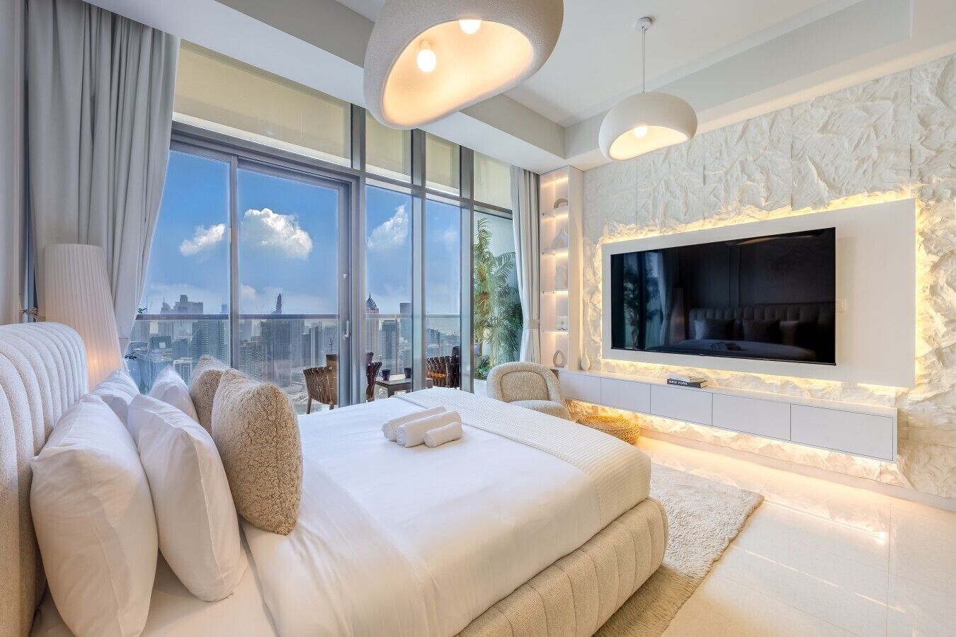 A modern hotel room with a large bed, a flat-screen TV mounted on a textured wall, and a city view through large windows.