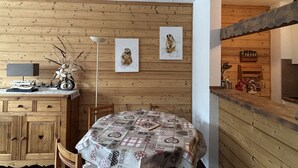 Een houten interieur met een eettafel, bedekt met een tafelkleed met een patroon, houten kasten, een lamp en twee aan de muur hangende kunstwerken.