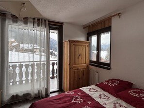 A bedroom with a bed, a wooden wardrobe, a window with a view of a snowy landscape, and a curtain. - Practical apartment, balcony - 5 guests (Châtel)