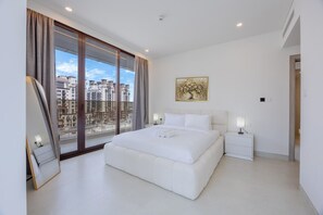 A modern bedroom with a large bed, a nightstand, a bedside table, a window with curtains, and a city view. - Minimalist 2 Bedroom l near Cricket Stadium (Dubai Sports City)