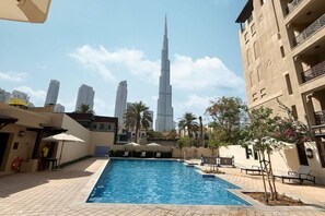 A swimming pool with lounge chairs and umbrellas, surrounded by buildings and palm trees, with a view of the Burj Khalifa in the background. - Central 1 Bed l Minutes Walk to Dubai Mall (Downtown)