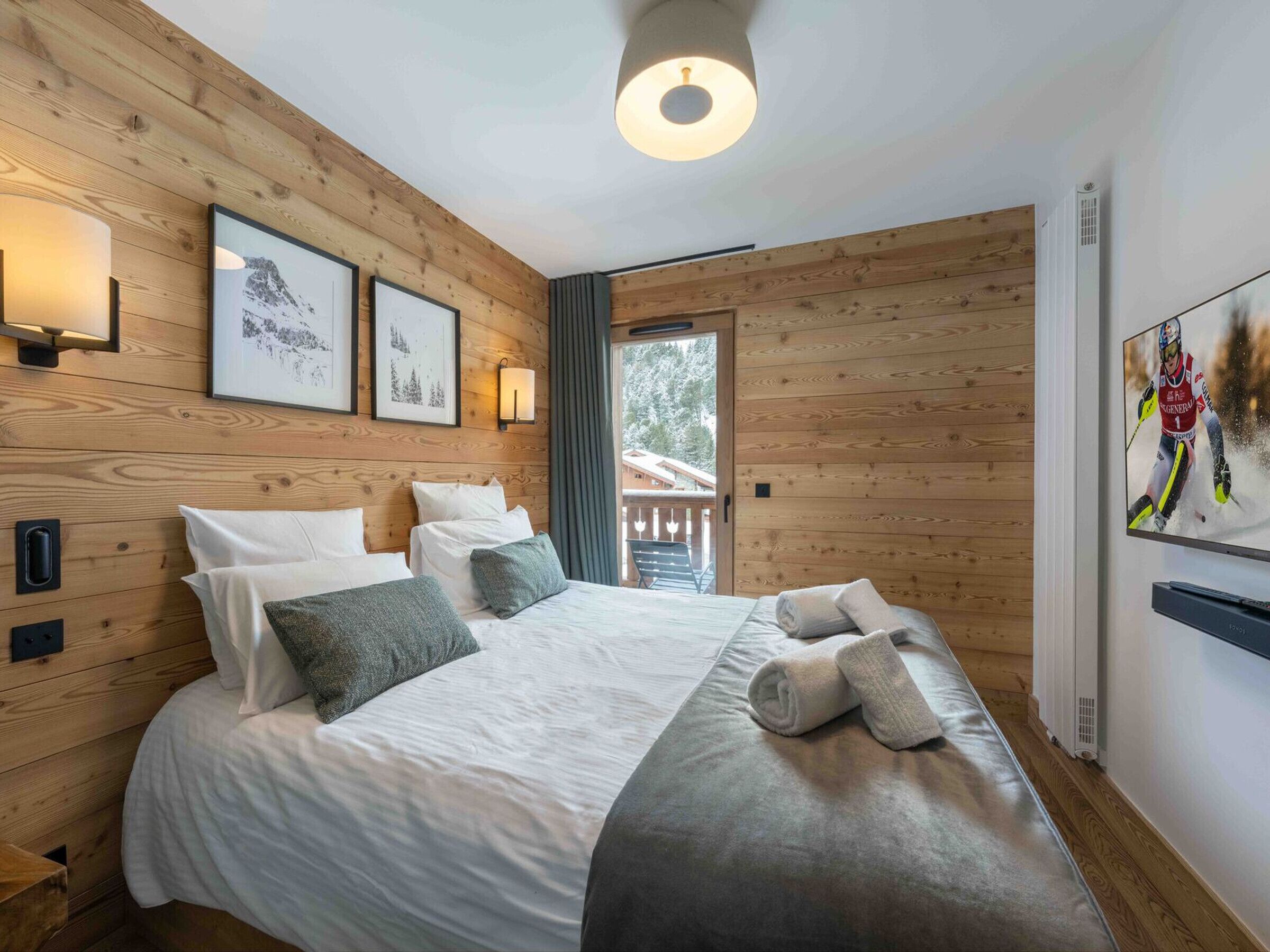 A hotel room with wooden panel walls, a bed with white linens, two wall-mounted lights, and a large window with a view of the outdoors.