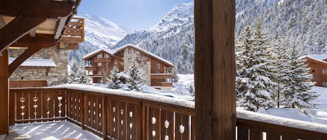 A wooden deck with a table and chairs, overlooking a snow-covered mountain landscape with buildings.