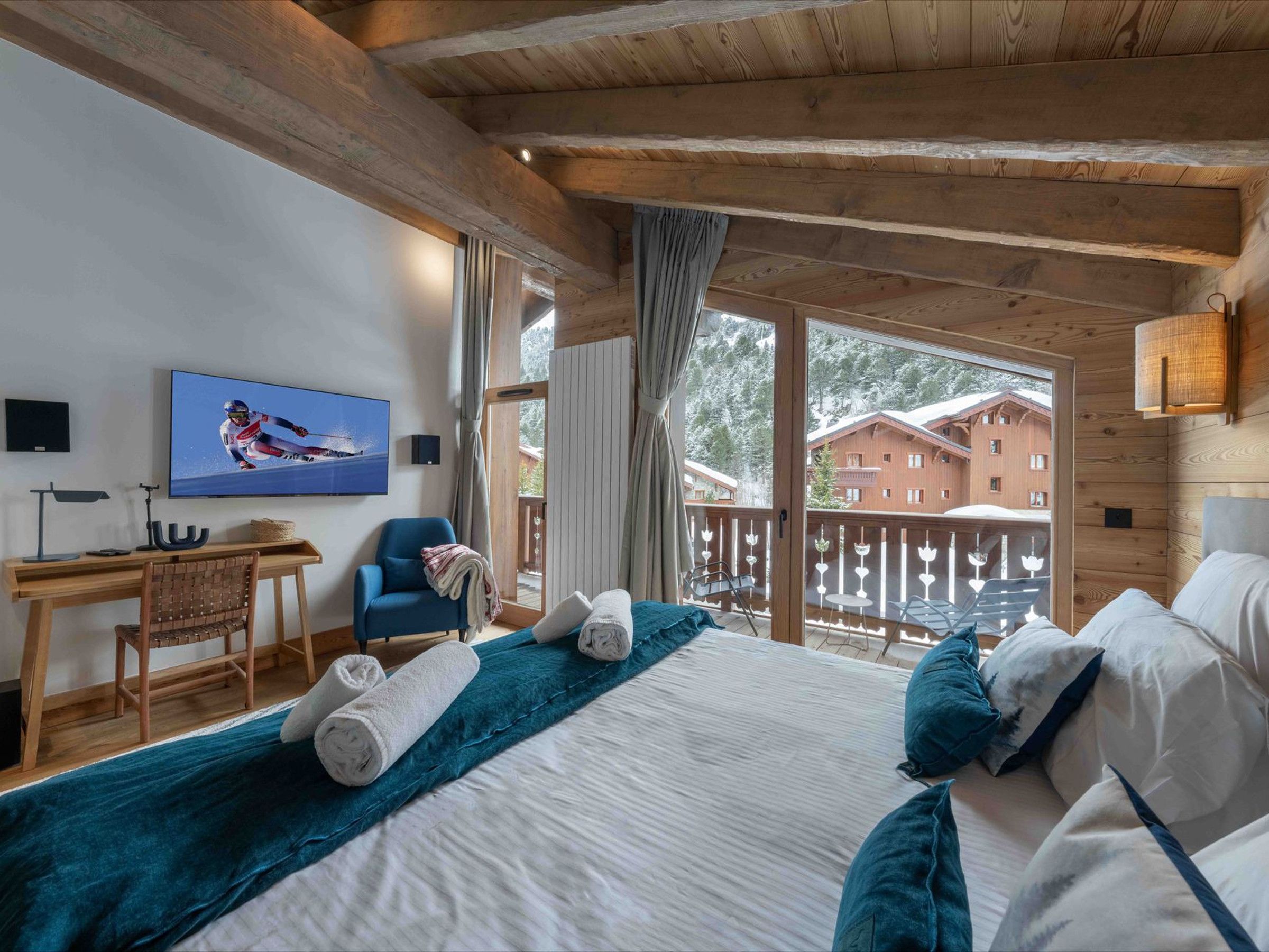 A wooden cabin room with a bed, a desk, a chair, a television, and a balcony with a view of buildings.