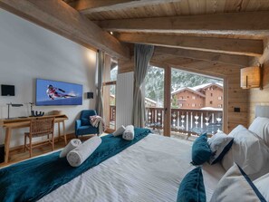 A wooden cabin room with a bed, a desk, a chair, a television, and a balcony with a view of buildings.
