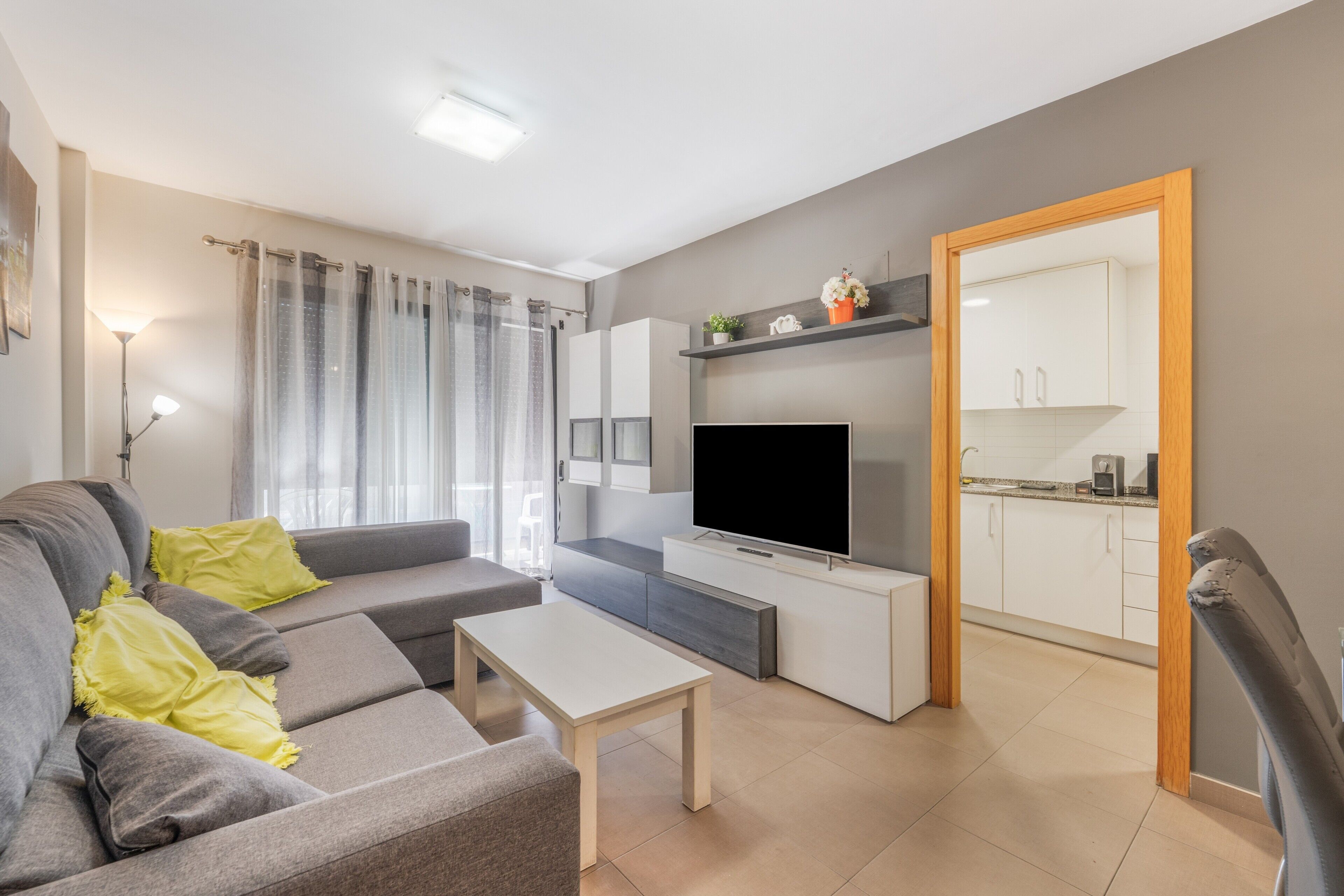 A modern living room with a sofa, a coffee table, a flat-screen TV, and a kitchen area visible through an open door.