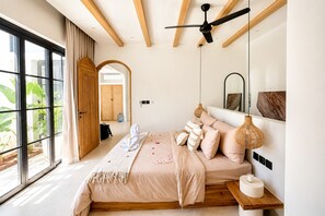 Un dormitorio moderno con un gran ventanal, un ventilador de techo, una cama con varias almohadas y una mesita de noche con lámpara.