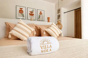Un lit bien fait, avec une literie beige, une serviette blanche ornée d’une broderie « VILLA SOLA », et trois œuvres d’art encadrées au-dessus du lit.
