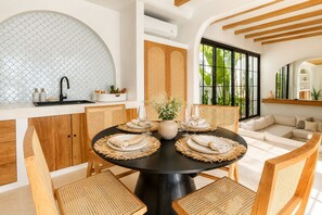Uma sala de jantar com uma mesa redonda posta para quatro pessoas, rodeada por cadeiras de vime, uma toalha de mesa preta e um vaso central.
