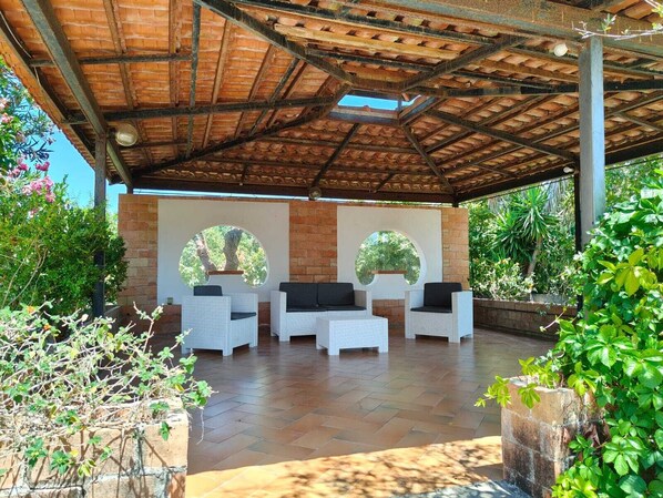 An outdoor patio with a brick wall, two round windows, and white wicker furniture. - A retreat for relaxation and nature. (Ascea)