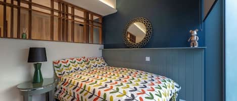 Un dormitorio con cama, mesita de noche con lámpara, espejo y un objeto decorativo en la pared.