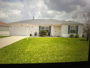 A single-story house with a front garage, a well-maintained lawn, and a small garden with a variety of plants. - 2-bedroom house in Lake Sumter The Villages includes Golf Cart (The Villages)