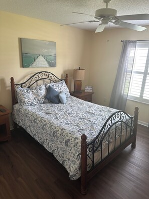 A bedroom with a bed, a nightstand, a lamp, a ceiling fan, a window with curtains, and a painting on the wall. - 2-bedroom house in Lake Sumter The Villages includes Golf Cart (The Villages)