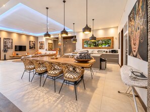 A modern kitchen with a dining area, featuring a large wooden table, wicker chairs, pendant lighting, and a large window with a view of greenery. - Spacious 4 BR villa Center Seminyak (Seminyak)