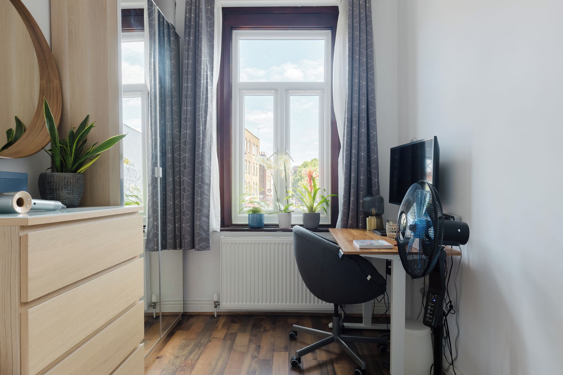 A modern office space with a desk, chair, and computer monitor. There is a window with curtains, a radiator, and a potted plant on the desk.
