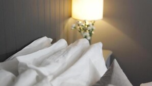 Uma cama com lençóis brancos e almofada cinza, um abajur e um vaso com flores em uma mesa de cabeceira.