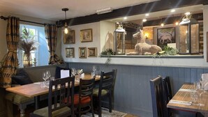 Interior de restaurante com mesas e cadeiras de madeira, uma estátua decorativa de cavalo, obras de arte emolduradas na parede e um teto com tonalidade azul.