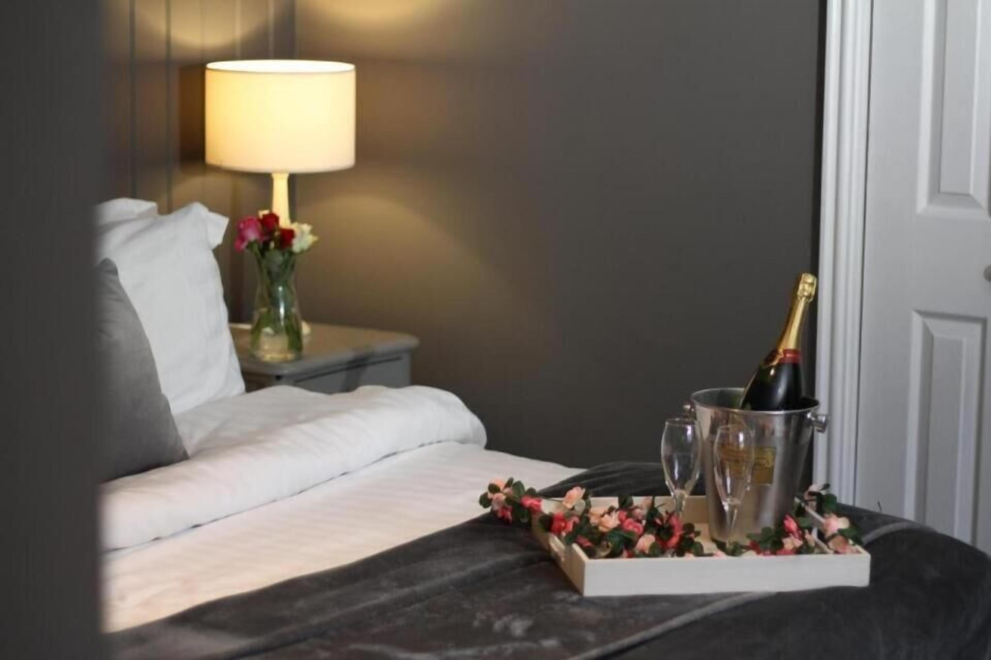 A neatly made bed with white linens, a bedside table with a lamp, a vase with flowers, and a tray with a bottle of champagne and glasses.