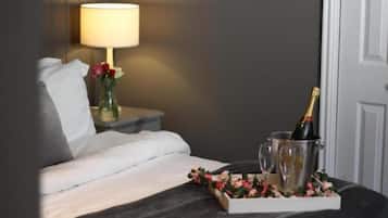 A neatly made bed with white linens, a bedside table with a lamp, a vase with flowers, and a tray with a bottle of champagne and glasses.