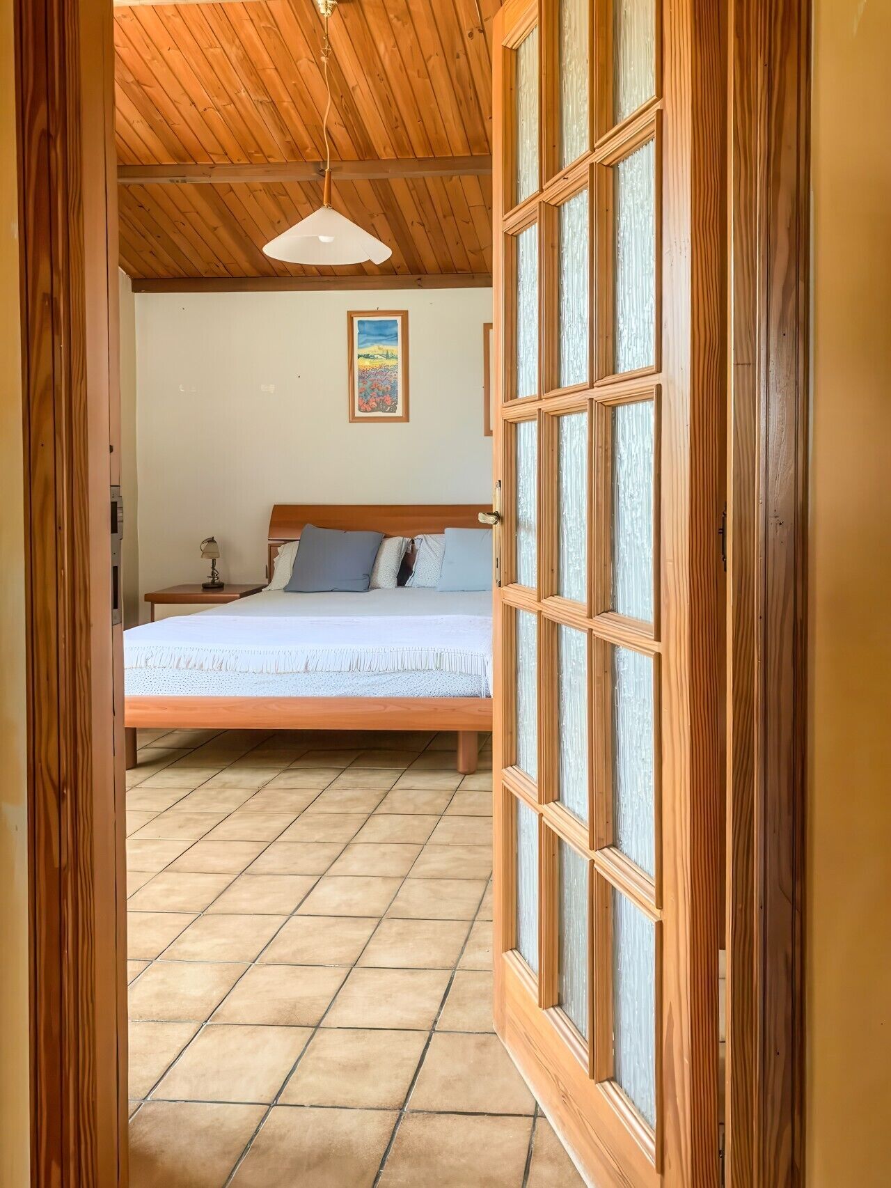 A bedroom with a wooden ceiling, a bed with a white bedspread, a painting on the wall, and a window with wooden frames.