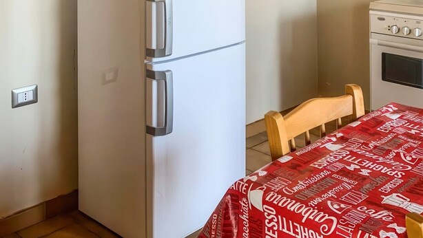 A kitchen with a white refrigerator, wooden cabinets, a table with a red tablecloth, and a wall-mounted oven. - Casa Paola - Iglesias (Iglesias)