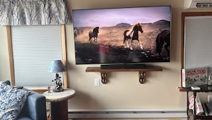 A living room with a mounted television displaying a horse scene, a sofa, a small table with a lamp, and a bookshelf with books and a Monopoly game. - Water Front 1 bedroom house on tranquil Smith Island (Smith Island)