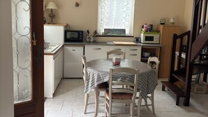 A kitchen with a dining table, chairs, a hanging light fixture, and a window with lace curtains. - Welcome to the mill (Épouville)