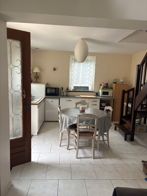A kitchen with a dining table, chairs, a hanging light fixture, and a window with lace curtains. - Welcome to the mill (Épouville)