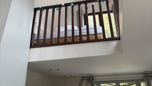 A room with a high ceiling, a wooden balcony railing, a bed with a patterned bedspread, and a window with curtains. - Welcome to the mill (Épouville)