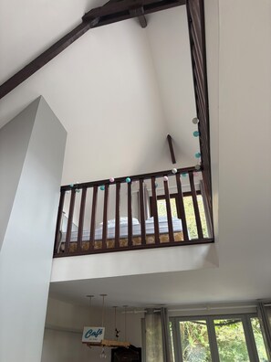 A room with a high ceiling, a wooden balcony railing, a bed with a patterned bedspread, and a window with curtains. - Welcome to the mill (Épouville)