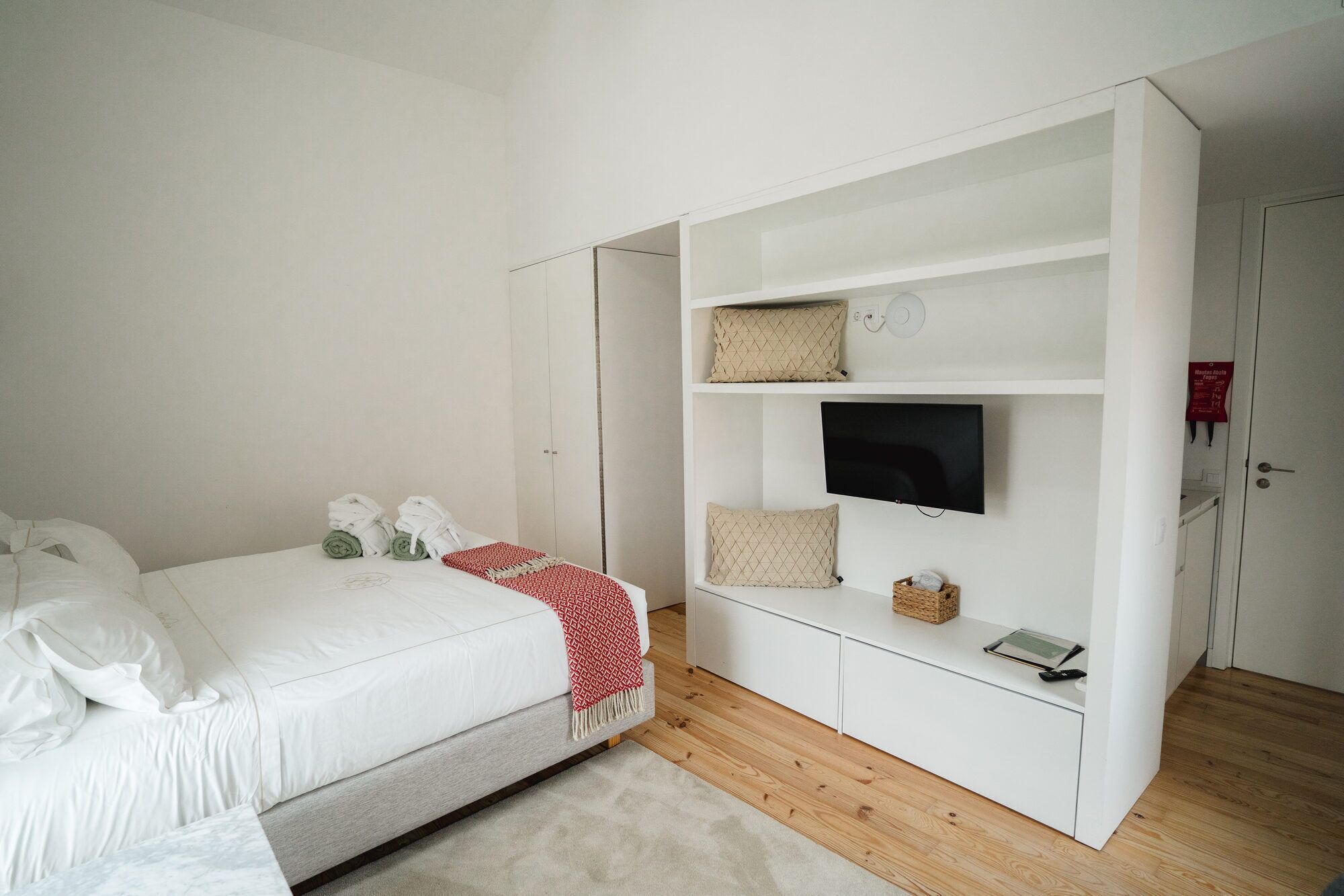 Un dormitorio con una cama, un televisor sobre un estante blanco y una puerta con un letrero rojo.