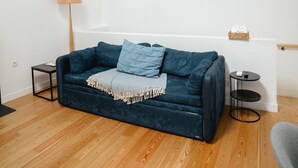 Ein modernes Wohnzimmer mit einem dunkelblauen Sofa, einem kleinen Beistelltisch, einer Lampe und einem Korb voller Gegenstände an einer weißen Wand.