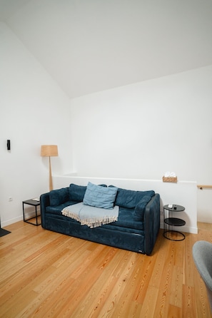 A modern living room with a dark blue sofa, a small side table, a lamp, and a basket of items on a white wall.